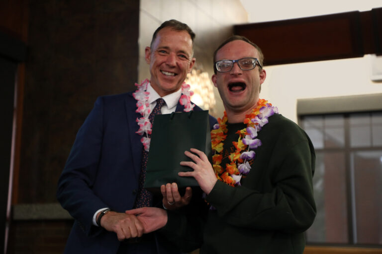 Two individuals smiling and celebrating, one presenting a gift while wearing a floral lei.